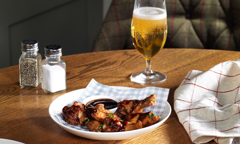 A table with Whitbread Inns Chicken Wings in a round dish with a pot of BBQ sauce, salt and pepper shakers and a small beer.