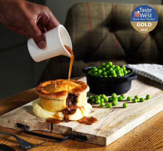 Someone pouring gravy onto a meat pie on top of mashed potato and a bowl of peas on a wooden board.